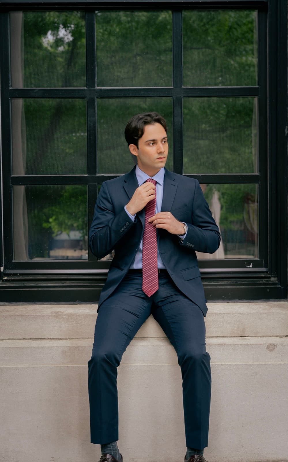 Gonzalo Pelenur seated on a stone ledge in a navy suit, adjusting a red tie